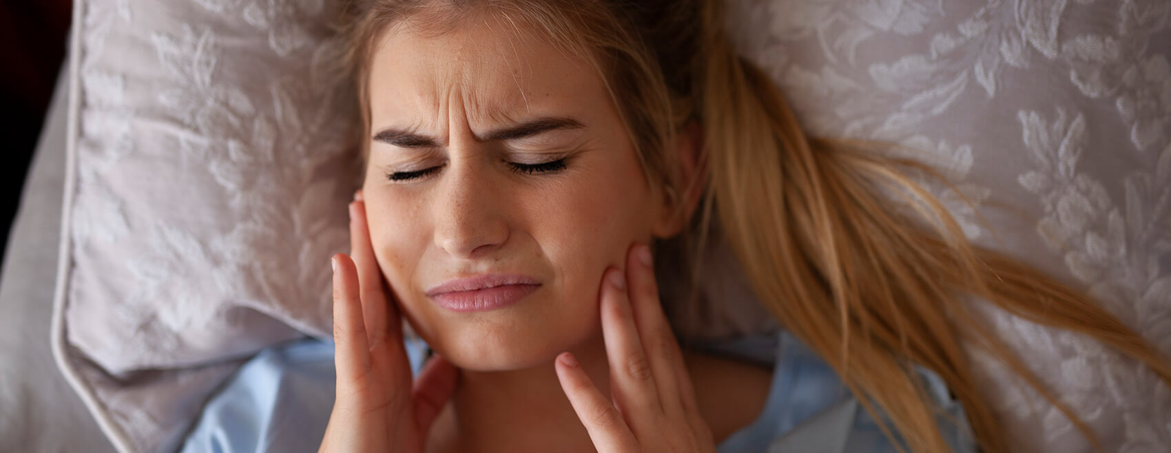 woman laying in bed holding both sides of her jaw in pain
