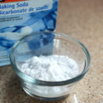A small glass bowl of white powder next to a blue box of baking soda